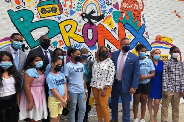 U.S. Education Secretary Miguel Cardona stands in front of a multi-colored mural of the word "Bronx" along with Schools Chancellor Meisha Porter and students and adults from PS/MS 5.